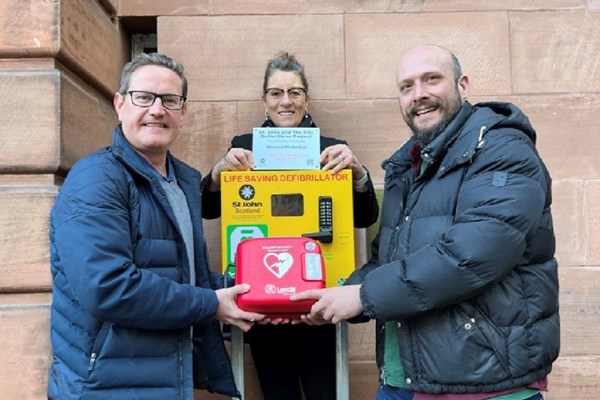 Cardiac arrest survivor Mike Pinkerton presents a defibrillator to the Church Hill Theatre, Edinburgh. St John Scotland volunteer Lynn Cleal holds the plaque. December 2021